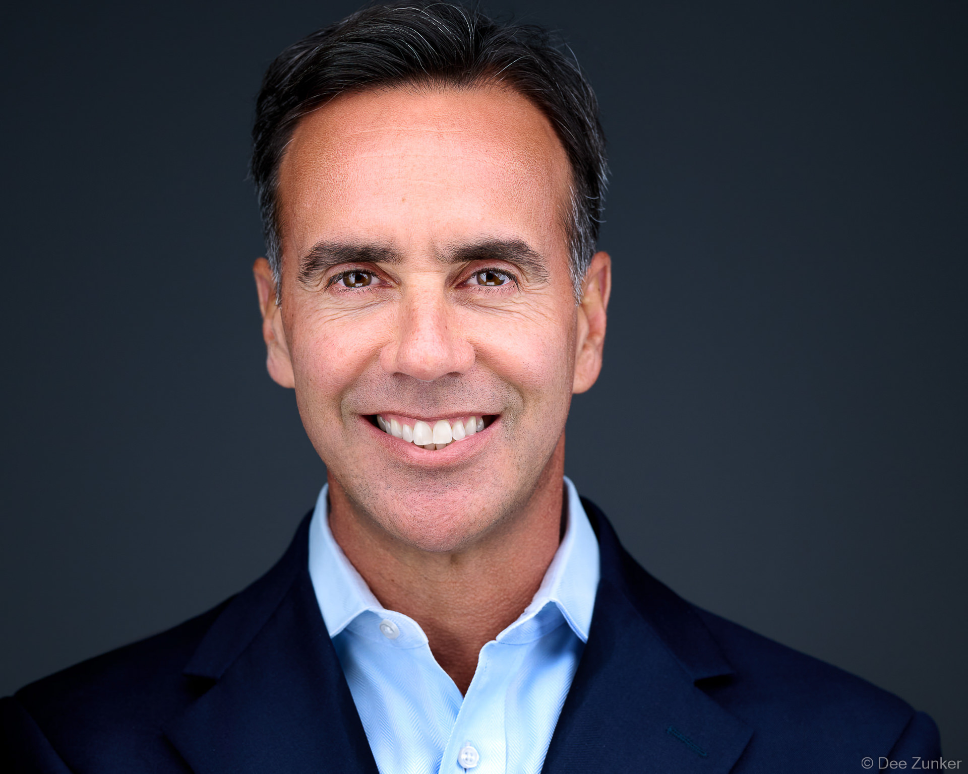 Male executive smiling in navy blazer and light blue shirt, professional studio headshot on dark grey backdrop, Bellaire Houston.