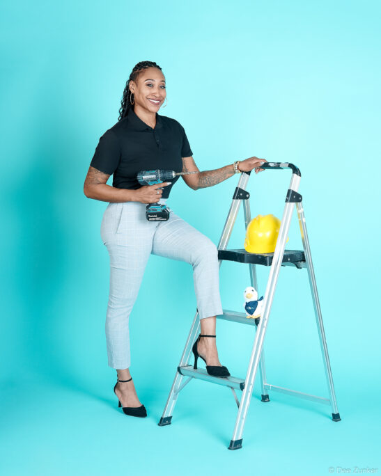 Houston branding portrait of female busines owner holding a Makita drill beside a ladder with a yellow hard hat and Aflac duck toy.