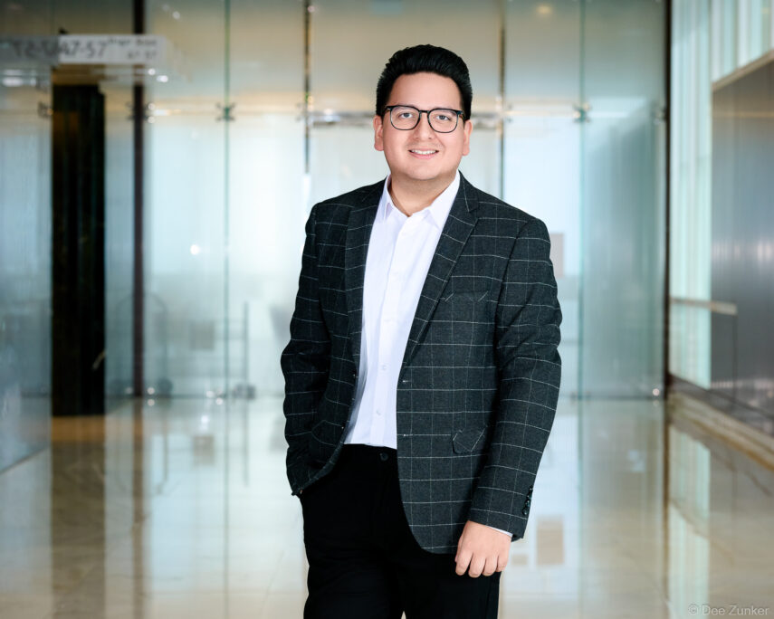 Professional male corporate headshot in dark plaid blazer and glasses, smiling in a modern Houston office lobby setting.