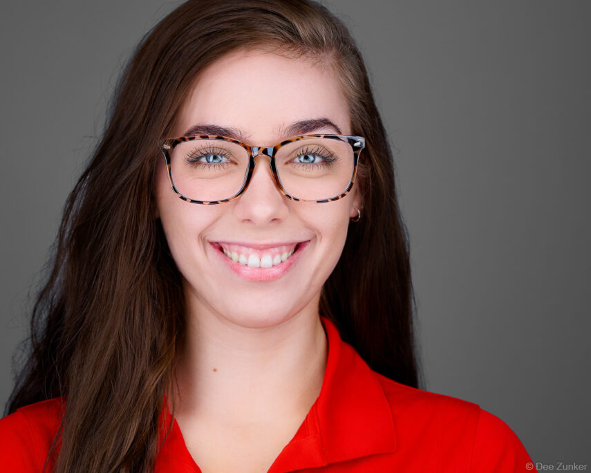 Smiling healthcare professional in red top with tortoiseshell glasses, studio corporate headshot on gray background, Houston TX.