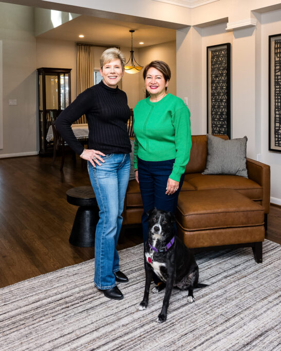 Two women and a black dog pose smiling in a modern Houston living room with leather sofa and hardwood floors.