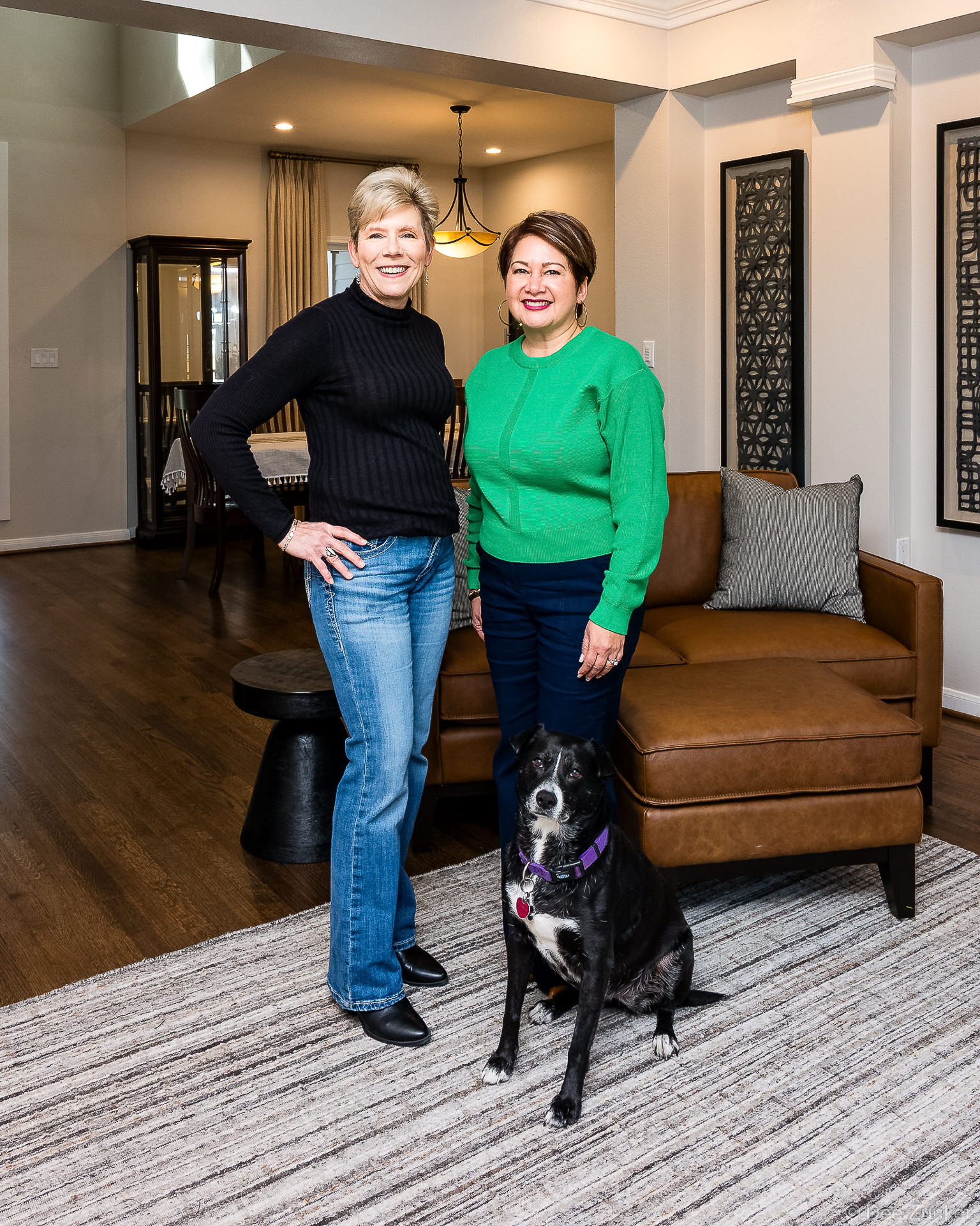 Two women and a black dog pose smiling in a modern Houston living room with leather sofa and hardwood floors.