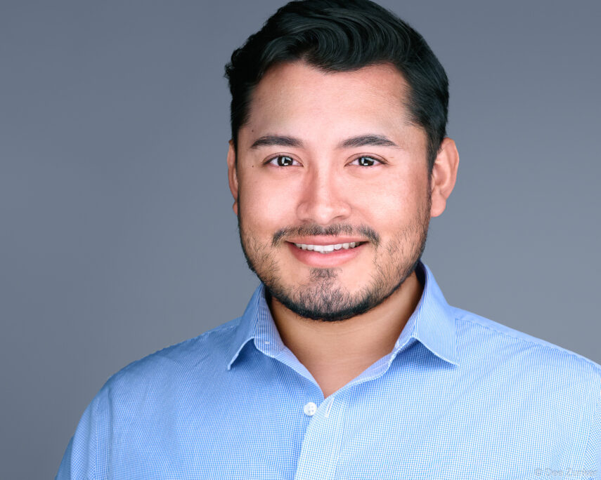 Professional corporate headshot of a male architect in a light blue shirt against a gray studio background, Houston, TX.