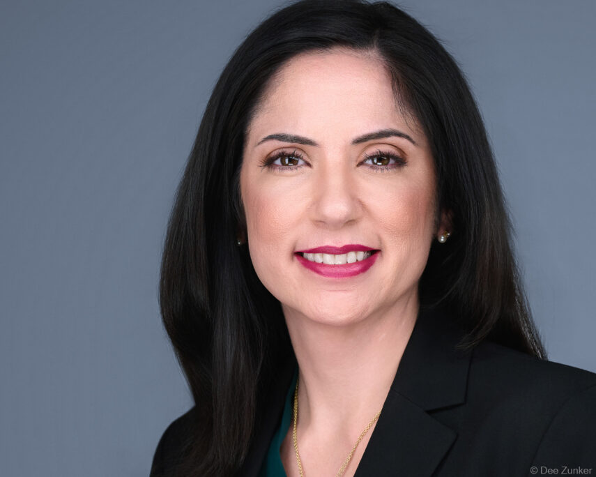 Professional corporate headshot of a Houston architect, smiling woman in black blazer with gray studio background, pearl earrings and gold necklace.
