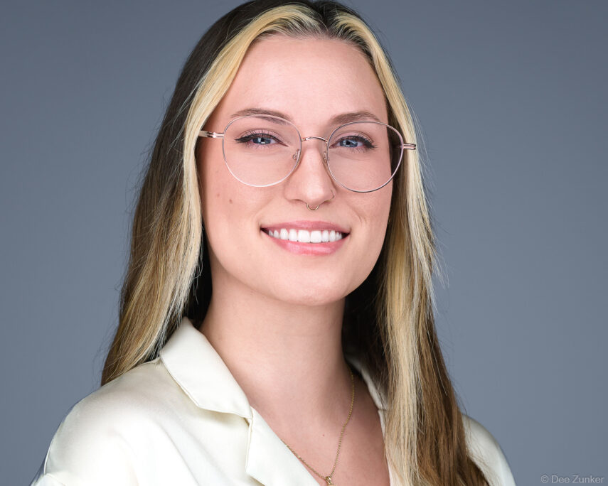 Female architect's corporate studio headshot, smiling, wearing glasses and cream blouse, photographed against a gray background in Houston.