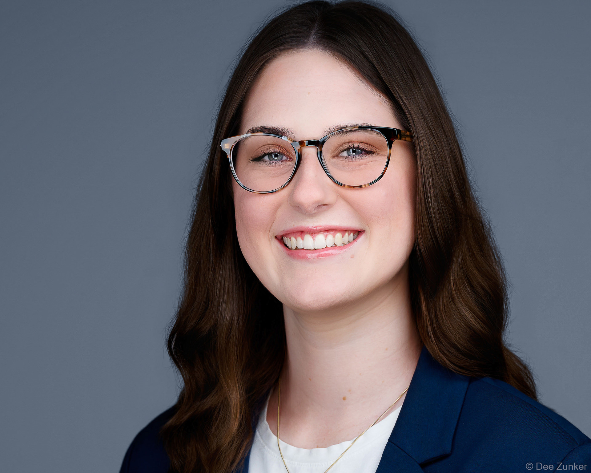 Professional corporate headshot of a female architect, smiling, wearing glasses and navy blazer, studio portrait with gray background, Houston TX.