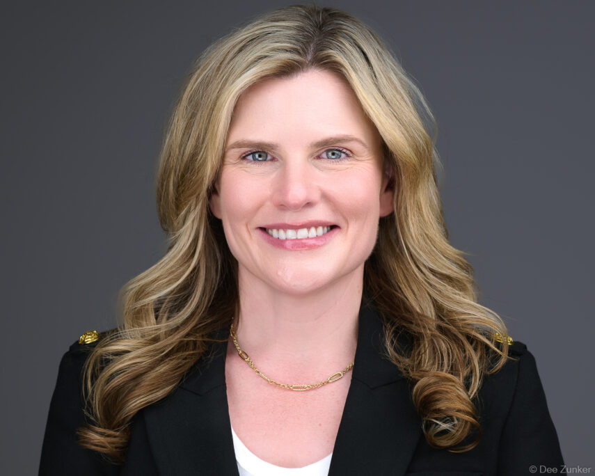 Professional corporate headshot of a smiling female attorney in a black blazer with gold necklace, photographed against a gray studio background in Houston.