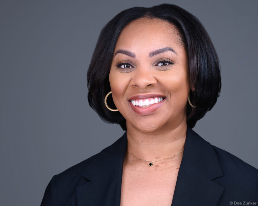 Professional corporate headshot of a Houston attorney, studio portrait with gray background, black blazer, and gold jewelry.