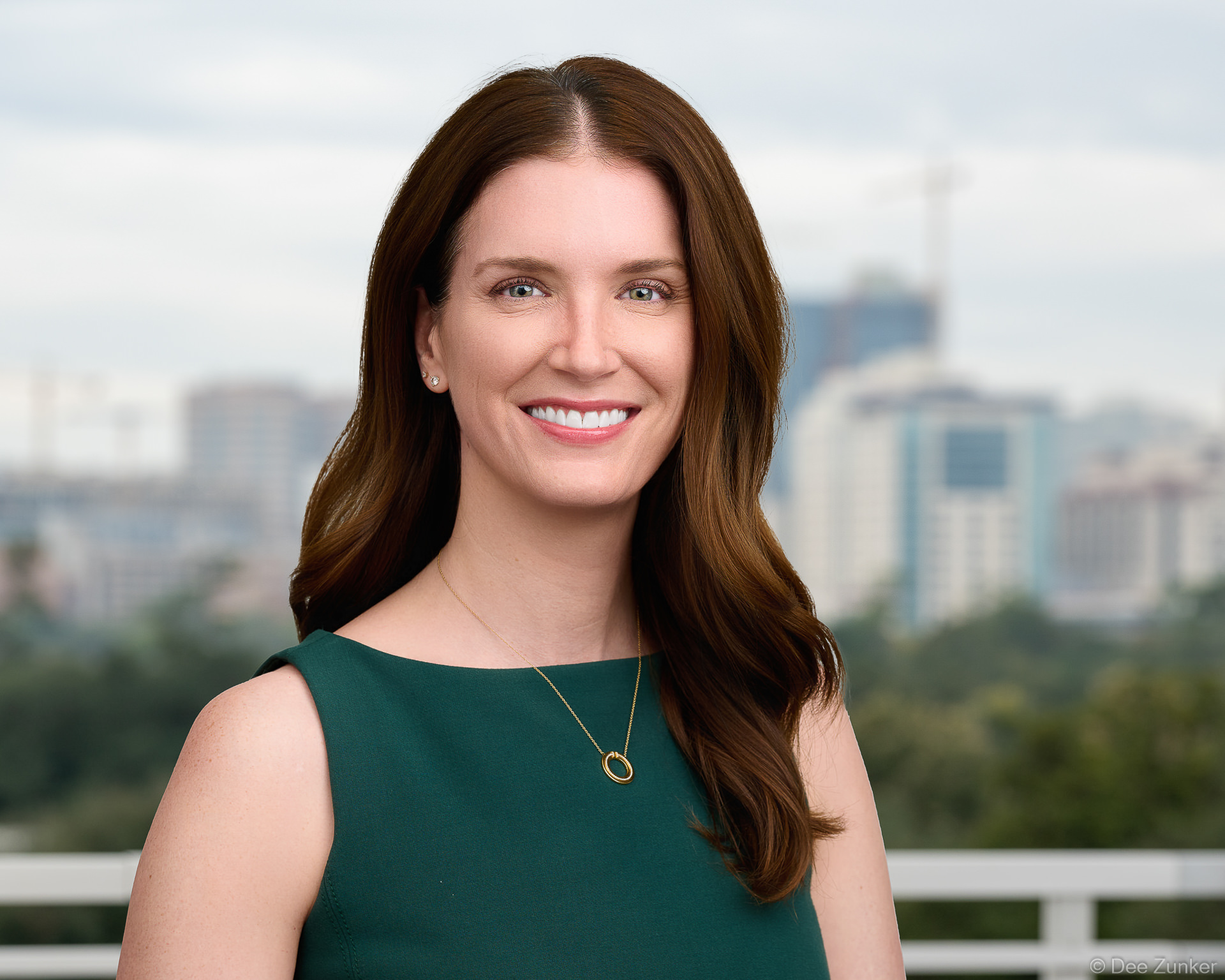 Professional environmental headshot of a female dermatologist or laser clinic physician, smiling outdoors against a blurred Houston cityscape background.