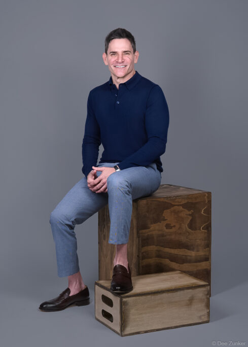 Man in navy polo and light gray trousers seated on wooden crate, smiling confidently in a Houston corporate bradning portrait by Dee Zunker Photography.