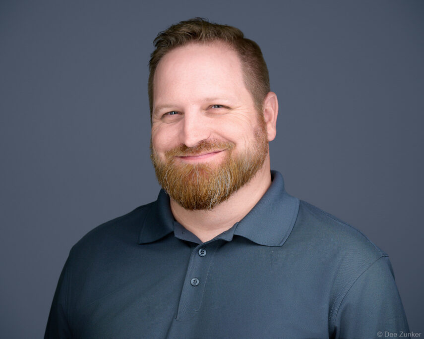 Professional studio headshot of a bearded male employee in a dark polo shirt, smiling against a gray background, Houston, TX.