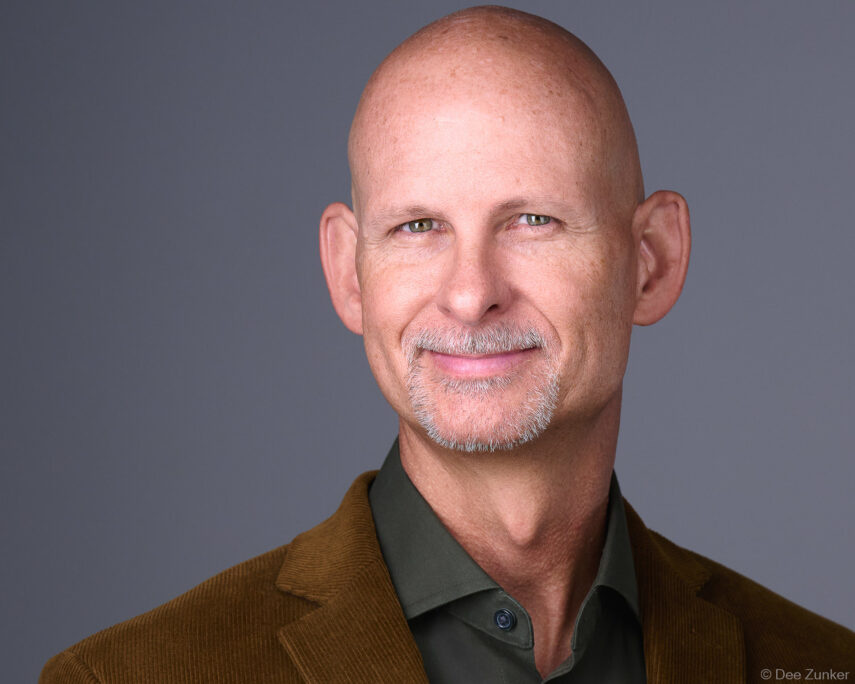 Bald male executive with gray goatee smiles in studio headshot, wearing brown corduroy blazer against gray background, Houston professional photography.