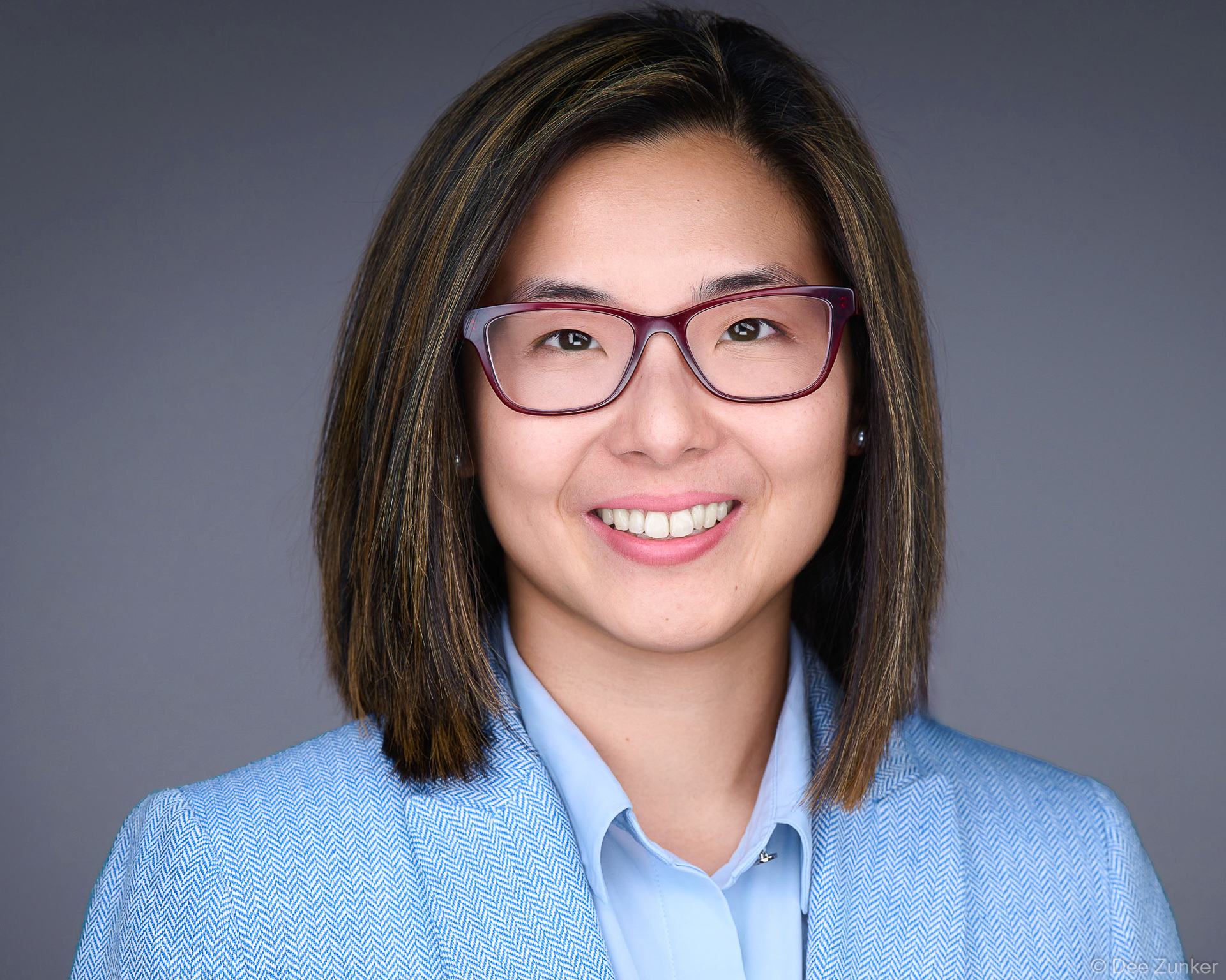 Professional woman in blue blazer and red glasses smiles in a studio corporate headshot against a gray background in Houston.