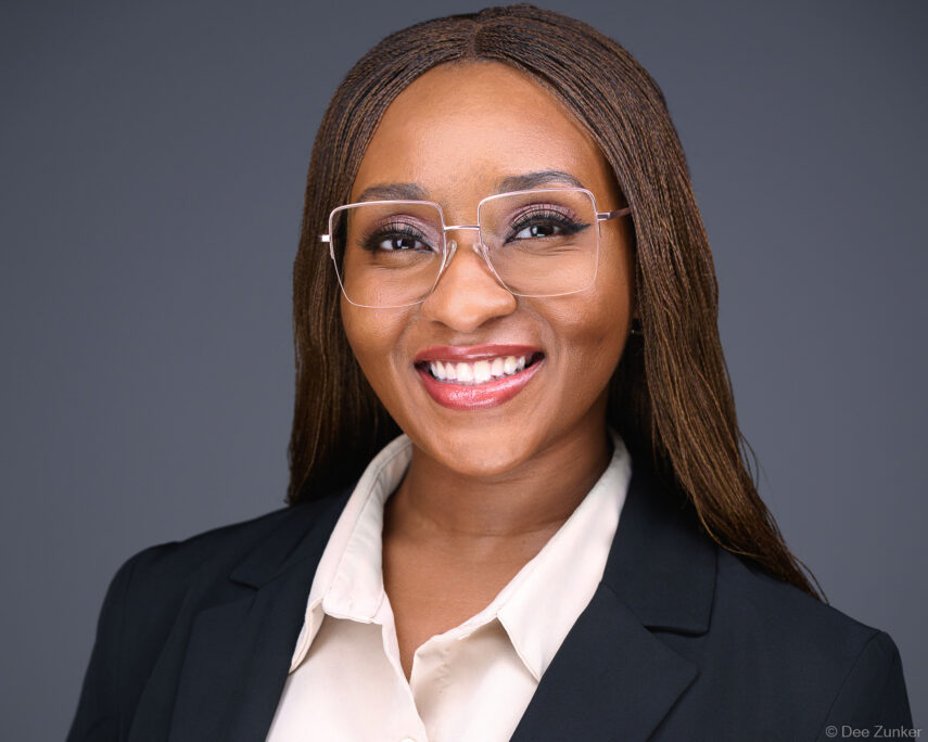 Professional woman in dark blazer and clear-frame glasses smiles in a Houston studio corporate headshot against a gray background.