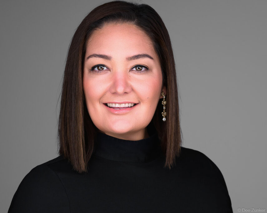 Professional woman's corporate headshot on gray background, dark turtleneck and gold earrings, studio portrait by Houston commercial photographer.