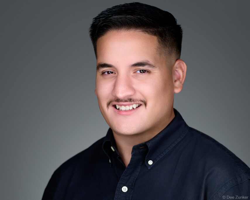 Young Hispanic male professional with mustache smiles in studio corporate headshot, wearing dark shirt against gray background, Houston TX.