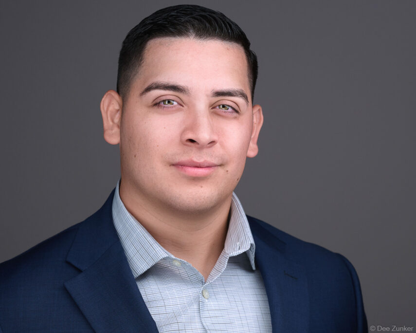 Professional corporate headshot of a Houston business professional in a navy blazer and checkered shirt against a gray studio background.