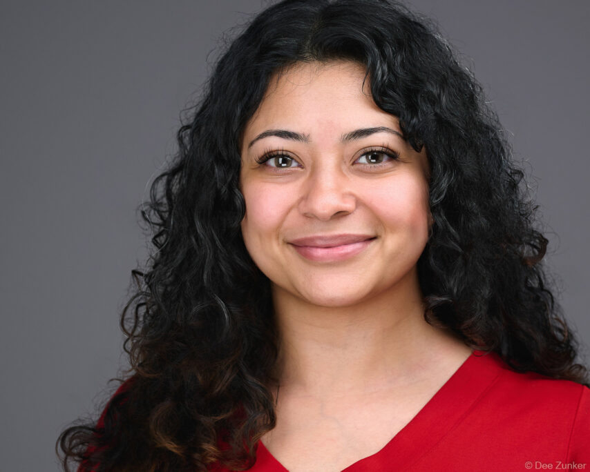 Professional corporate headshot of a woman with curly dark hair wearing a red top against a gray studio background in Houston.