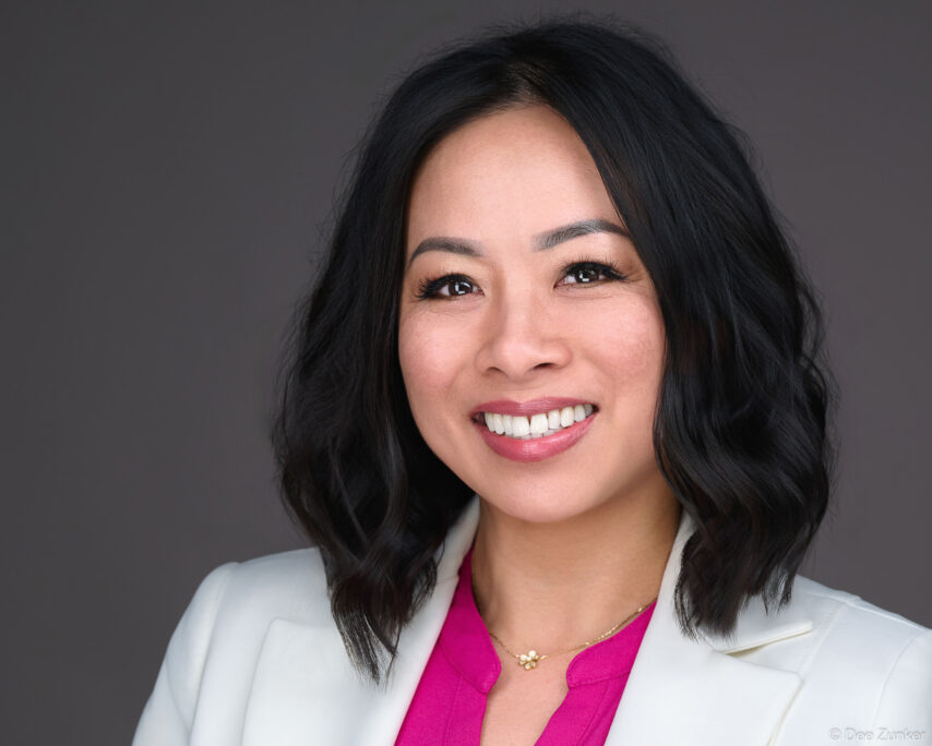 Professional Houston corporate headshot of a smiling woman in a white blazer and pink top against a gray studio background.