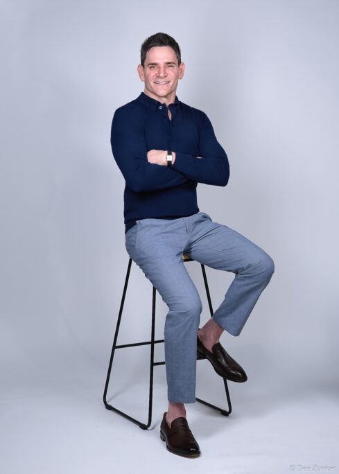 Man in navy polo and gray trousers sits cross-armed on barstool, smiling, in a Houston corporate headshot by Dee Zunker Photography.