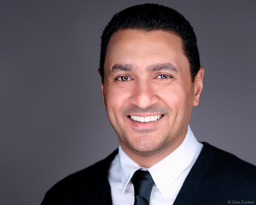 Studio corporate headshot of a smiling male professional in a dark suit and black tie against a gray background, Houston, TX.