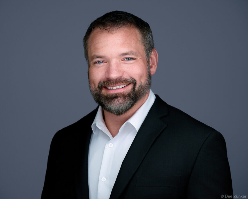 Male drilling industry executive smiles in studio corporate headshot, wearing black blazer and white shirt against gray background, Houston TX.