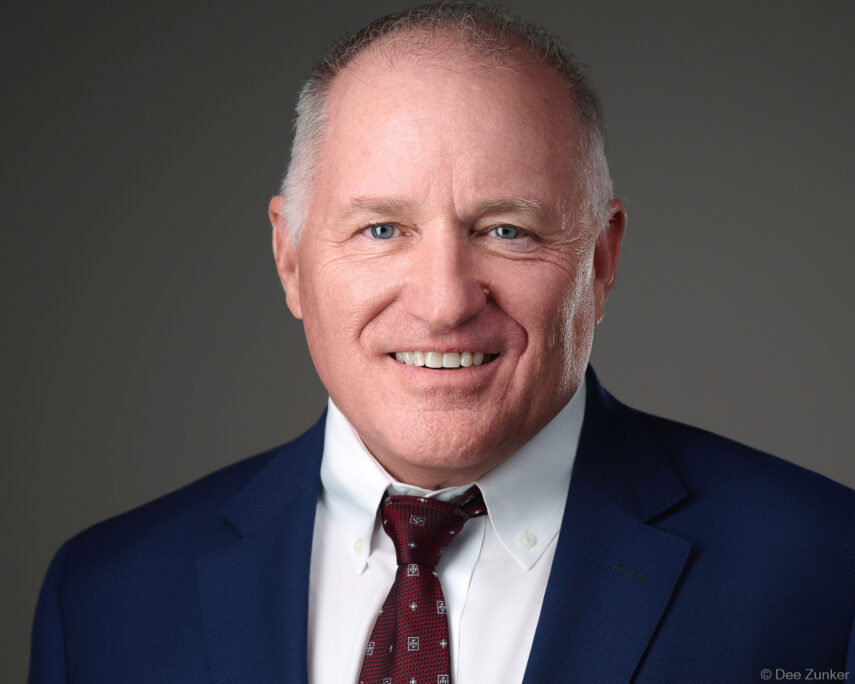 Professional corporate headshot of a senior male executive in a blue suit and burgundy tie, shot on a gray studio background in Houston.