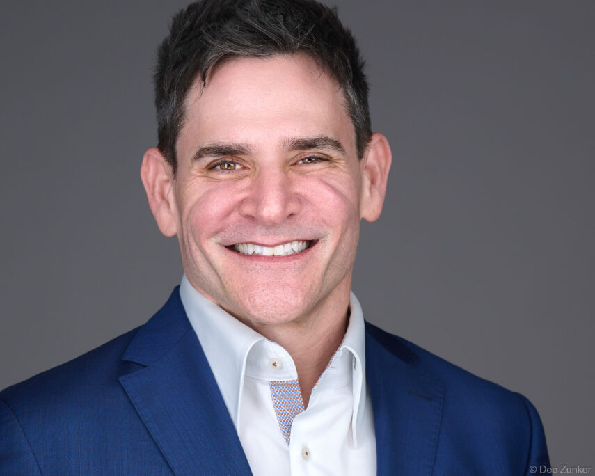 Male executive smiles in a navy blazer and white shirt against a gray studio background in a Houston corporate headshot.