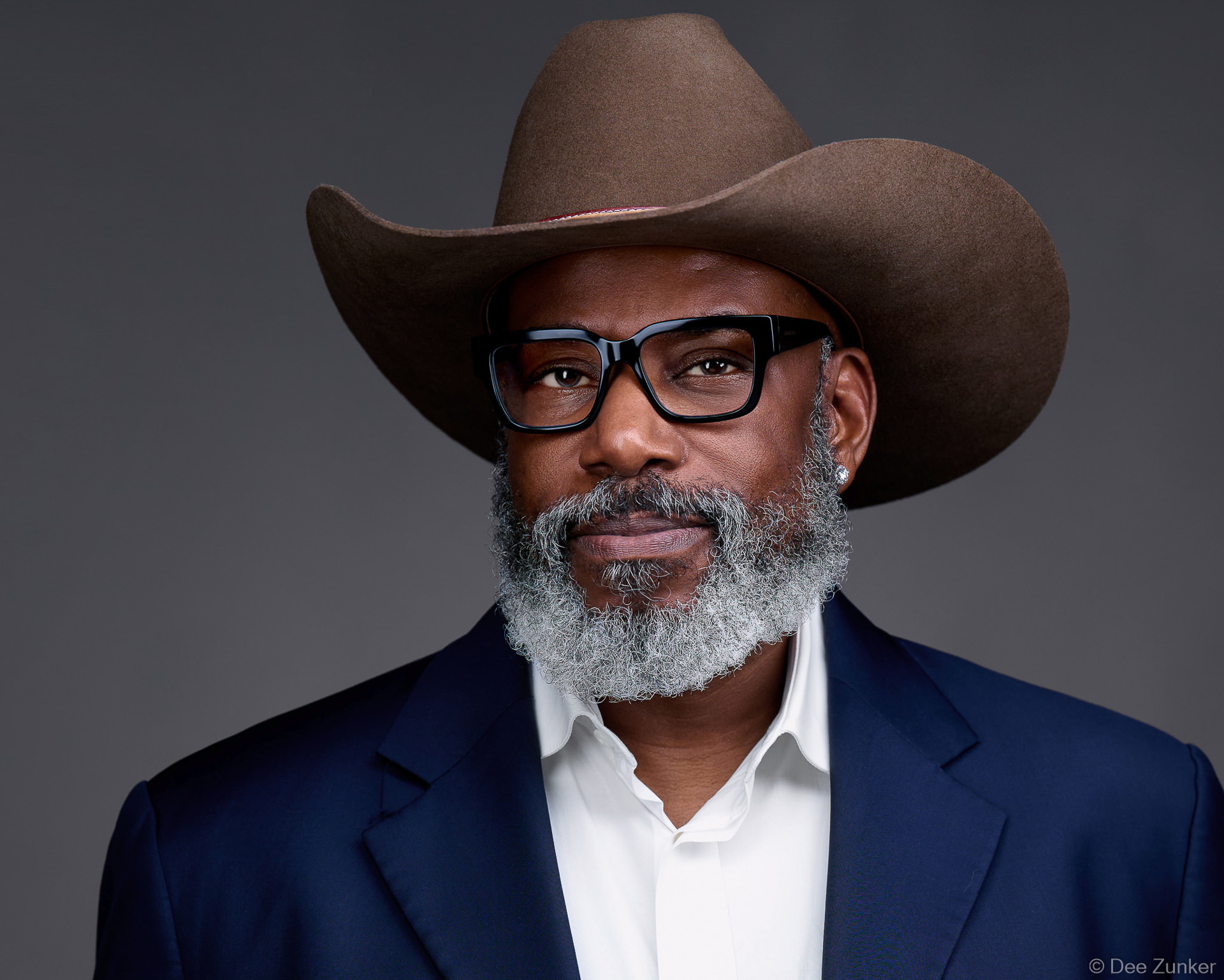 Professional studio headshot of a Houston executive man wearing a brown cowboy hat, black glasses, and navy blazer against a gray background.