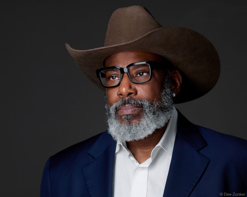 Professional studio headshot of a man with a gray beard, cowboy hat, glasses, and navy blazer, shot against a gray background in Houston.