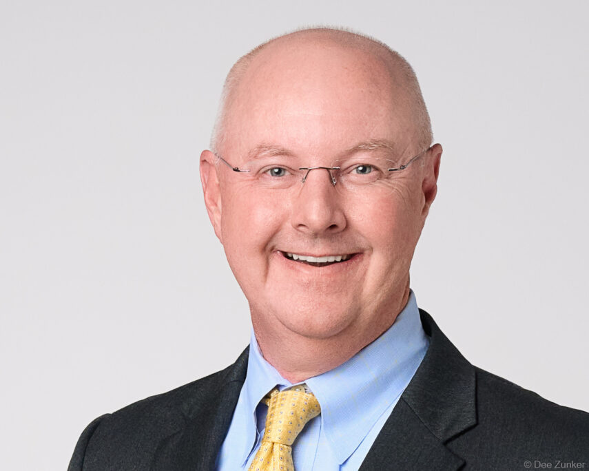 Studio corporate headshot of a smiling male financial executive wearing glasses, light blue shirt, and gold tie against a gray background, Houston TX.