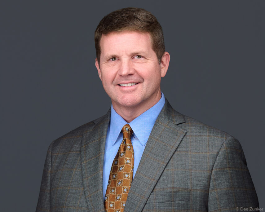 Professional corporate headshot of a Houston law firm attorney in a gray suit and gold tie, studio portrait with gray background.