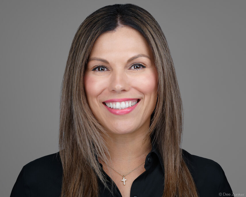 Female attorney at a Houston law firm smiles in a studio headshot against a gray background, wearing a black blazer and cross necklace.