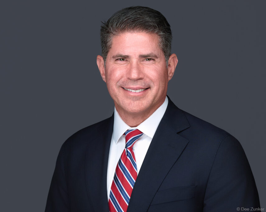 Professional corporate headshot of a male Houston law firm executive in a navy suit and striped tie against a gray studio background.