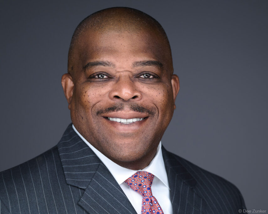 Male physician in pinstripe suit and patterned tie smiles confidently in a Houston studio headshot against a gray background.