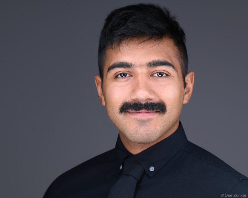Houston professional corporate headshot of a young man with a mustache wearing a black shirt and tie against a gray studio background