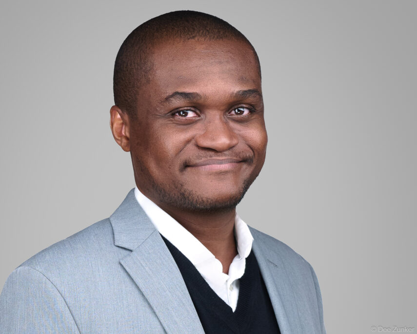 Male dermatology professional in light gray blazer and black sweater smiles in a studio corporate headshot, gray background, Houston TX.