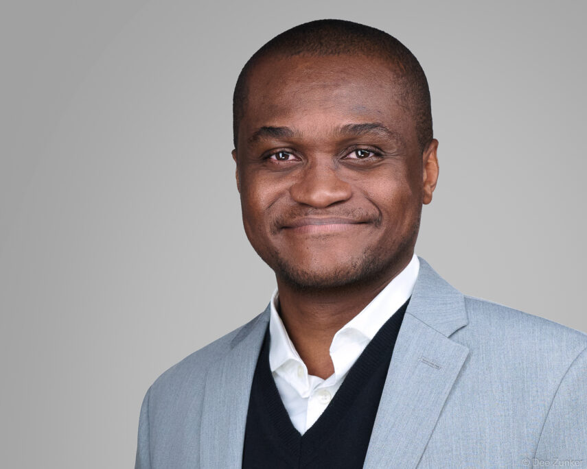 Male dermatologist in gray blazer and dark sweater smiles warmly in a professional studio headshot against a gray background, Houston TX.
