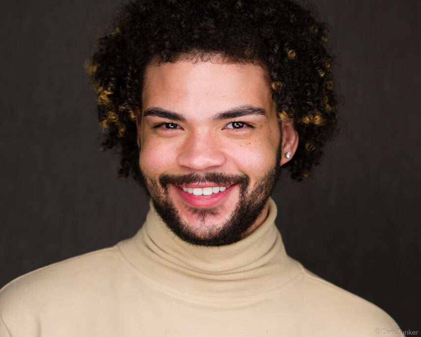 Young professional man with curly hair and beard smiles in a studio corporate headshot, wearing a beige turtleneck, dark background, Houston.