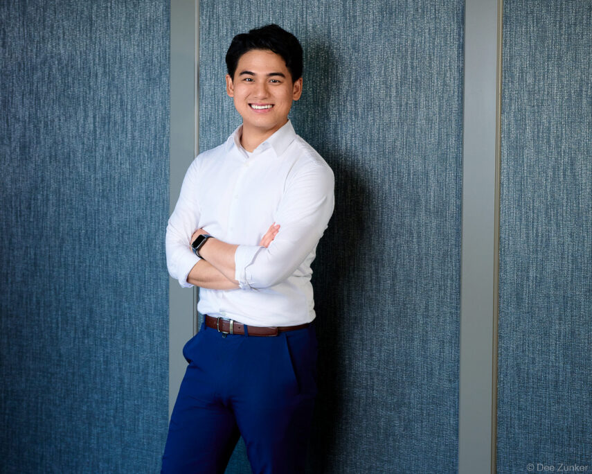 Smiling young male professional in white dress shirt and blue trousers, arms crossed, leaning against blue textured wall, Houston corporate headshot.