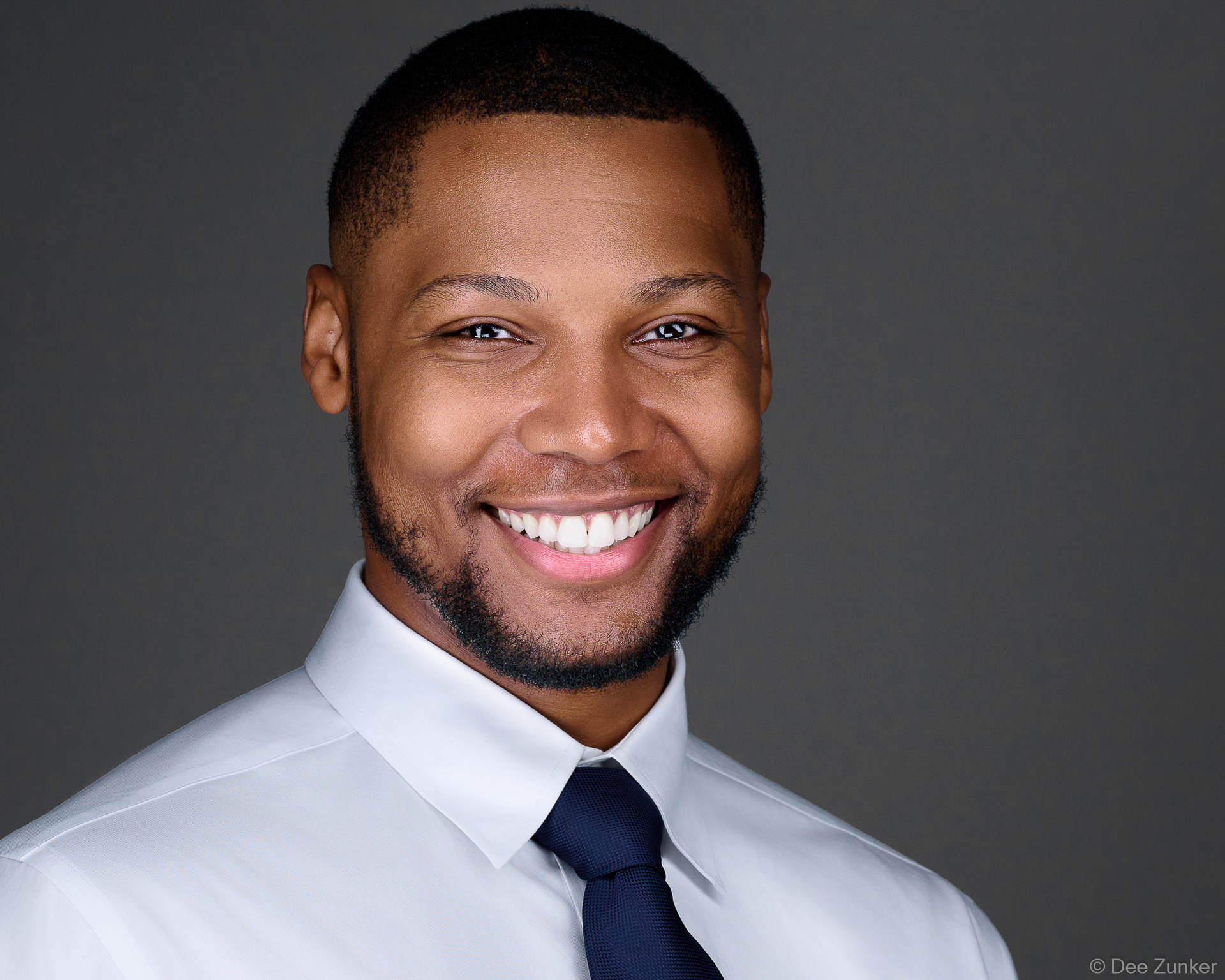 Professional corporate headshot of a smiling man in a white dress shirt and navy tie against a gray studio background, Houston TX