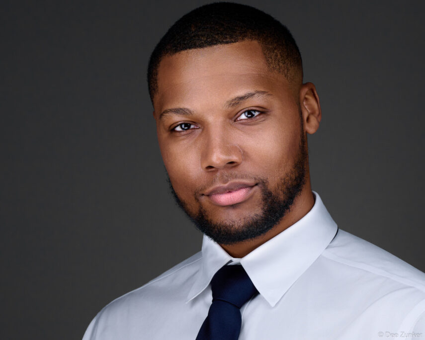 Professional corporate headshot of a man in a white dress shirt and navy tie against a gray studio background, Houston TX.
