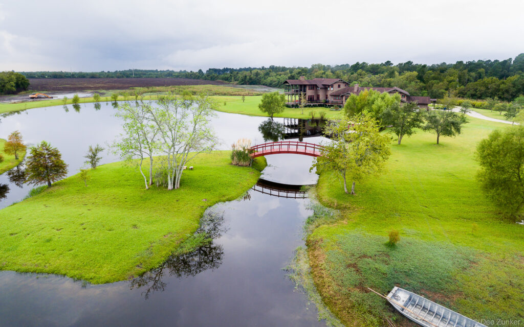 Drone aerial photo of Jamison Lake estate in Sealy TX featuring a red arched bridge, pond channels, and large lodge surrounded by green grounds.