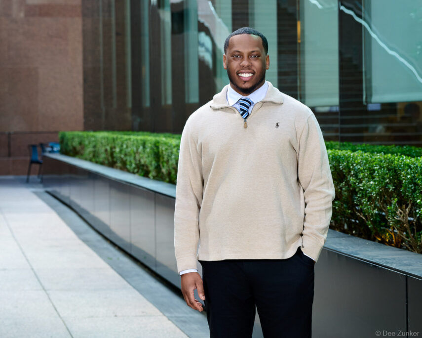 Smiling man in beige Polo quarter-zip and striped tie poses outdoors for a Houston corporate headshot, modern office building backdrop.