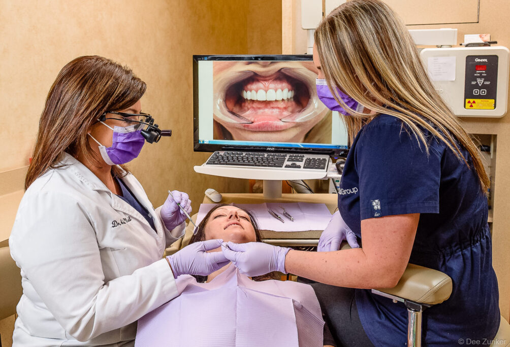 Female dentist and assistant treating a patient in a modern dental office, with intraoral camera imagery displayed on monitor