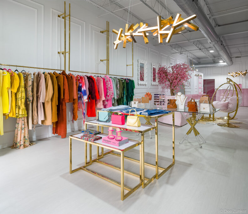 Colorful clothing racks, gold display tables with handbags and shoes, and decorative cherry blossom tree inside Lilly's Kloset Houston boutique.