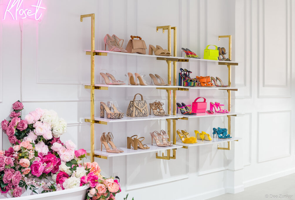 Gold wall-mounted shelves displaying colorful heels and handbags at Lilly's Kloset Houston boutique, with pink floral arrangement and neon sign.