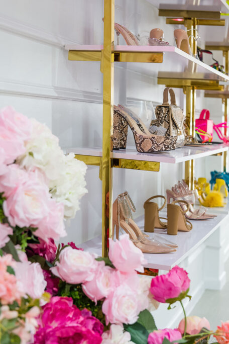 Gold shelving unit displaying heeled sandals and snakeskin platform shoes at Lilly's Kloset Houston boutique, with pink floral arrangement in foreground.