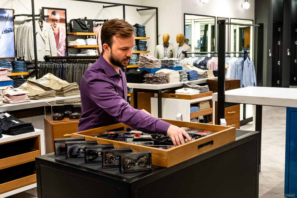 Male retail associate in purple shirt organizing boxed bow ties at a menswear store display counter, Houston commercial branding photography.