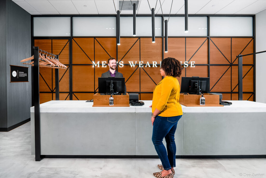 Female customer at Men's Wearhouse checkout counter with smiling male associate behind modern wood and concrete cash wrap desk.